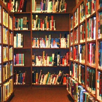 A library aisle with wooden bookshelves filled with books on both sides and at the end. The shelves are neatly organized, holding a variety of colorful books. The aisle is well-lit, creating a warm and inviting atmosphere for reading and studying, supported by scholarship granting organizations.