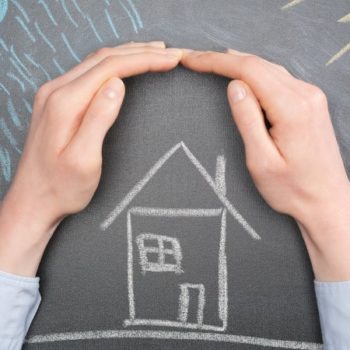 A pair of hands form a protective gesture over a chalk drawing of a house on a blackboard. The simple house, complete with door, window, and chimney, is surrounded by blue lines representing rain and yellow lines suggesting sunlight. This image evokes the warmth and security that charitable deductions can help provide to those in need.