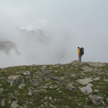 A person in a yellow jacket carrying a large backpack stands on a rocky and grassy terrain, looking out into the distance where heavy fog obscures the view of distant mountains.