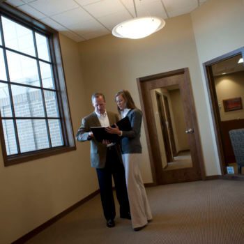 A man and a woman are standing in an office hallway, looking at a document together. The man is wearing a blazer and the woman is in business casual attire. They are near a large window and a closed door, with office furniture visible in the background.