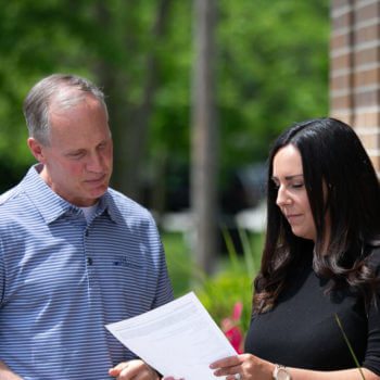 Two individuals stand outside, engaged in a discussion. The man on the left wears a striped polo shirt, while the woman on the right, clad in a black top, holds a document. They both examine it attentively; it appears to be related to powers of attorney. The background is lush and green.