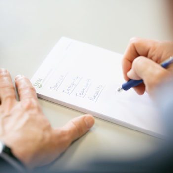 A person's hand is seen writing on a notepad with a blue pen. The notepad has a list of four words: "Ideas," "Information," "Teamwork," and "Results." The person is writing in a well-lit space with a blurred background.