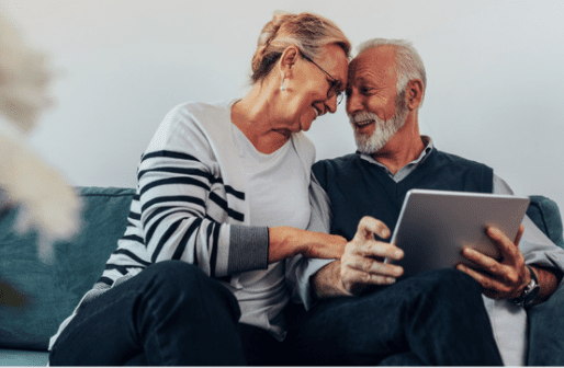 An elderly couple sits closely on a teal sofa, smiling and looking at a tablet together. The woman, wearing glasses and a striped sweater, leans towards the man, who is wearing a dark vest over a shirt. Both appear happy and engaged, perhaps perusing the services options of their trusted advisory firm.