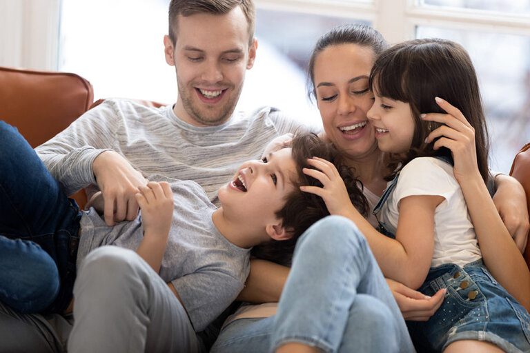 A family of four sits closely on a couch. The parents smile while their two children, a boy and a girl, laugh and playfully interact. Following their gifting guidelines, the family appears relaxed and happy, enjoying their time together in a cozy indoor setting.