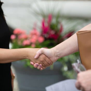 Two people shaking hands in an outdoor setting. Both individuals are shown from the wrist down. The left person, with nails painted purple, is wearing a black sleeve, while the right person is holding a brown bag and some papers related to disability insurance. Flowers are blurred in the background.