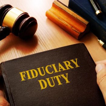 A person holds a book titled "FIDUCIARY DUTY" in gold letters. In the background, there is a judge's gavel, a wooden block, and another book on a wooden surface, signifying the legal responsibilities associated with managing group retirement plans.