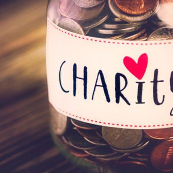 A glass jar filled with coins and a few bills, labeled "Charity" with a red heart above the letter "i", stands on a wooden table—a humble testament to charitable giving.