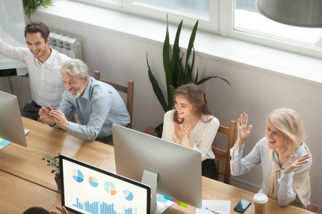 Four colleagues, three women and one man, are sitting at a long office table working on computers. They are smiling and raising their hands in celebration or applause. The setting is a bright office with large windows, depicting charts on computer screens related to elder care.