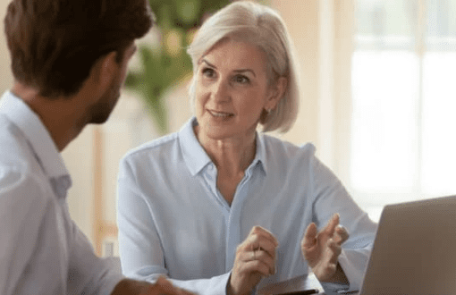 An older woman with short gray hair, dressed in a light blue blouse, is sitting at a table with a laptop in front of her, engaging in a conversation with a younger man who is seated facing her. The setting appears to be the casual office of an advisory firm.