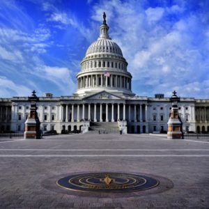 The image shows the United States Capitol building in Washington D.C., featuring its prominent white dome, columns, and majestic architecture. The foreground includes a circular emblem embedded in the pavement, with a backdrop of a partly cloudy blue sky—symbolic of safeguarding retirement savings for future generations.