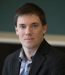 Shane Bradley, a man with short, dark hair wearing a blazer over a checked shirt, looks directly at the camera. The background is out of focus, placing emphasis on his face and attire.