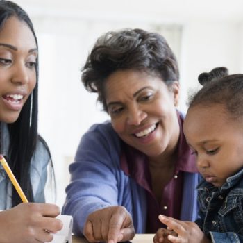 A young woman holding a pencil interacts with an elderly woman and a toddler. The elderly woman is smiling and pointing at something on the table, perhaps discussing the benefits of a 529 College Savings Plan, while the curious toddler looks engaged. They are sitting together in a bright, cozy setting.