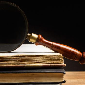 A magnifying glass with a wooden handle rests on top of a stack of books with aged, yellowed pages, placed against a dark background. The scene is softly lit, highlighting the texture of the book covers and the glass, evoking an atmosphere reminiscent of thorough fiduciary investigation.