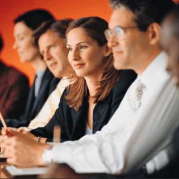 A diverse group of professionals sits in a row at a conference or meeting. They are wearing business attire and are attentively listening, taking notes, or looking forward. The background is a vibrant orange color.
