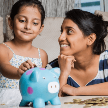 A young girl and a woman smile as the girl places a coin into a blue piggy bank. The woman, resting her chin on her hand, looks on proudly. They are seated at a table cluttered with more coins. The surroundings suggest a cozy indoor setting.
