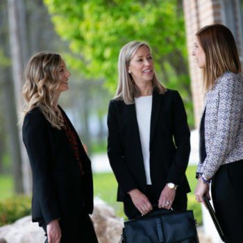 Three women in business attire are standing outdoors, engaged in conversation. They are smiling and appear to be in a professional setting. The background is blurred with green foliage and a building visible.