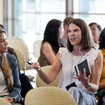 A woman in a white sleeveless top is engaged in conversation, gesturing with her hands and holding a notebook and pen. She is seated among other people in a conference or meeting setting, with light-filled windows in the background.