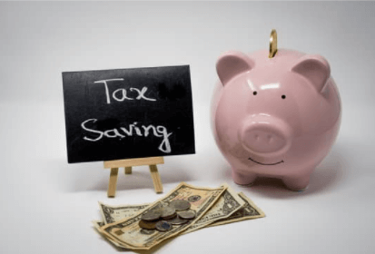 A pink piggy bank stands next to a small blackboard on an easel that reads "Tax Saving." There are various denominations of paper currency and a few coins placed in front of the piggy bank and blackboard, all on a plain white background.