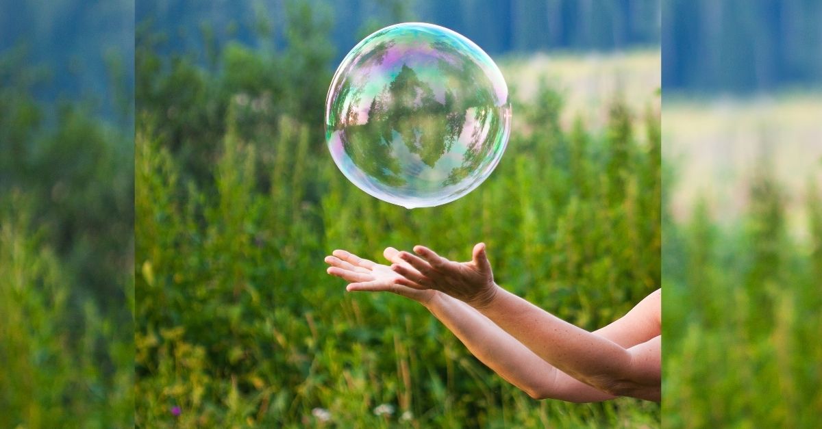 A large, iridescent bubble floats above a person's outstretched hands in a lush green outdoor setting. The background is filled with blurred foliage, emphasizing the bubble in mid-air.