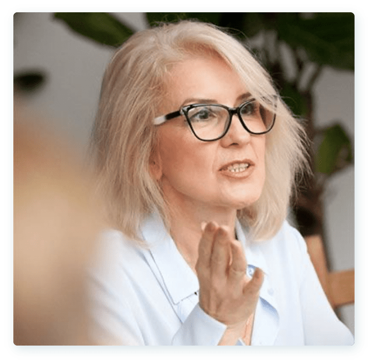 A woman with shoulder-length blonde hair and black-framed glasses is speaking. She is wearing a light blue blouse and gesturing with her right hand. The background shows blurred foliage.