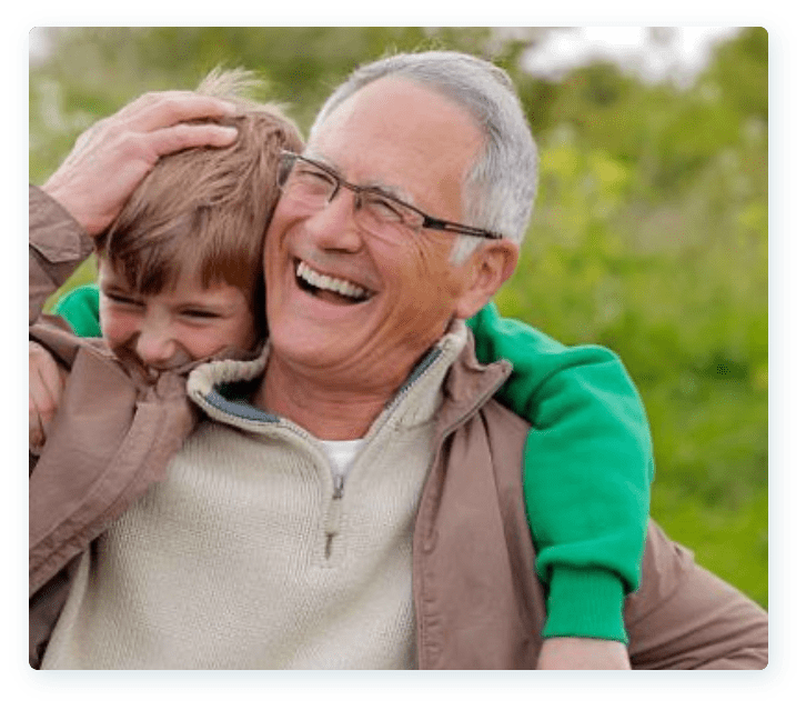 An elderly man with gray hair and glasses laughs heartily while holding a smiling young boy in a green sweater on his shoulders. They are outdoors, with a blurred background of greenery. The older man has his hand affectionately on the boy's head.