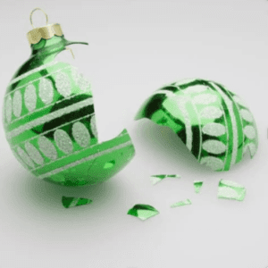 A shattered green Christmas ornament lies on a plain surface. The ornament, decorated with white and silver stripes and patterns, is broken into several pieces, with the top part still partially intact and the rest fragmented into small shards.