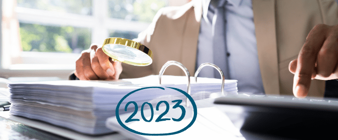 A person in a suit examines a stack of documents with a magnifying glass, while using a calculator. The year "2023" is written in blue and circled at the bottom of the image, possibly highlighting the new 2023 IRS limits. Bright, natural light fills the room from a window in the background.