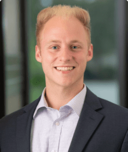 A young man with blond hair, wearing a light blue checkered shirt and a dark blazer, smiles at the camera. The background is blurred, showcasing what appears to be a window with a green outdoor view.