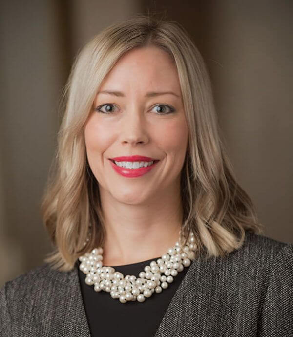 A woman with shoulder-length blonde hair smiles at the camera. She is wearing a dark grey blazer over a black top and a chunky pearl necklace. The background is blurred.