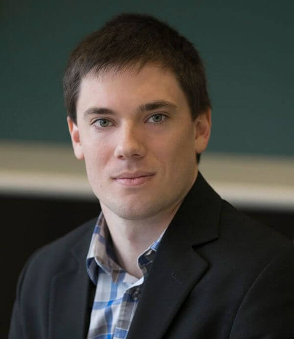 A person with short dark hair and fair skin is wearing a dark blazer over a checked shirt. They are looking directly at the camera with a neutral expression. The background is blurred and consists of green and dark colors.