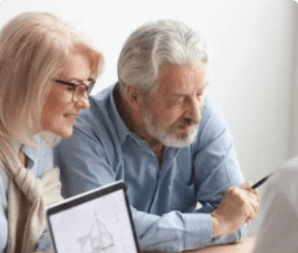 An elderly couple, both wearing glasses and blue shirts, are sitting together. The man is holding a pen and looking at a document, while the woman is smiling and looking at a computer screen that displays a sketch.