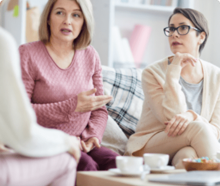 Two women are sitting on a couch engaged in conversation. One woman is wearing a pink sweater and gesturing with her hand, while the other woman, wearing glasses and a cream-colored sweater, listens attentively. A table with tea cups and a plate of snacks is in front of them.