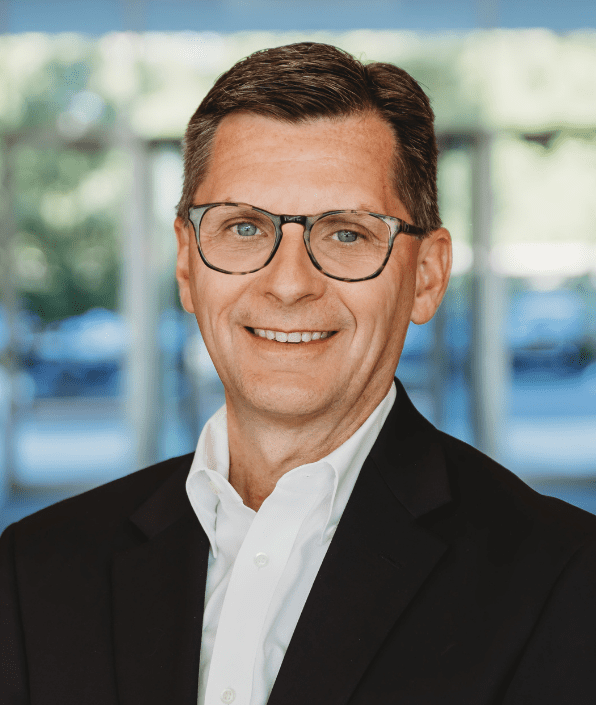 A middle-aged man with short brown hair and wearing glasses is smiling. He is dressed in a black blazer over a white shirt. The background is blurred, showing an indoor setting with large windows and some greenery outside.