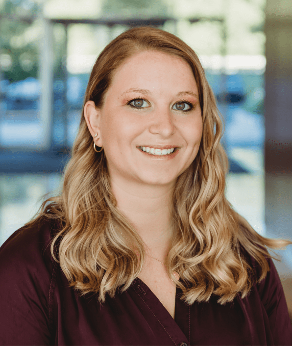 A woman with shoulder-length wavy blonde hair smiles at the camera. She is wearing a dark burgundy blouse and has a natural makeup look. The background is blurred, featuring a mix of indoor and outdoor elements with soft lighting.