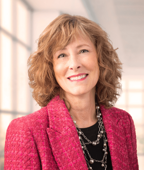 A woman with shoulder-length wavy brown hair smiles warmly at the camera. She is wearing a pink blazer and a multi-strand necklace. The background is softly lit, with large windows allowing natural light to fill the space.