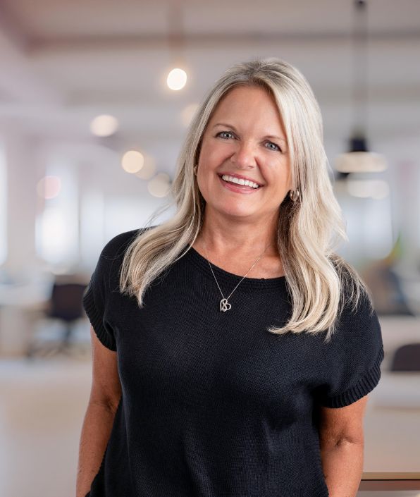 A mid-aged woman standing in an office room with blonde hair and blue eyes in a black shirt