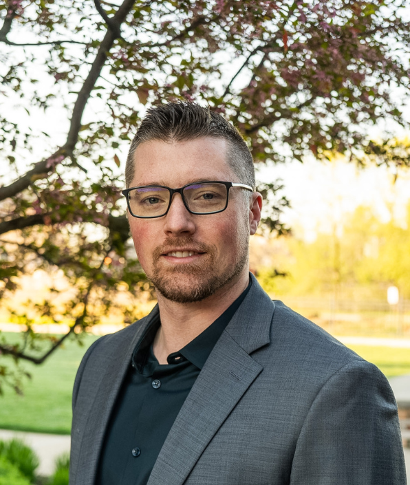 A young man with a goatee and glasses in a suit standing outside under a tree