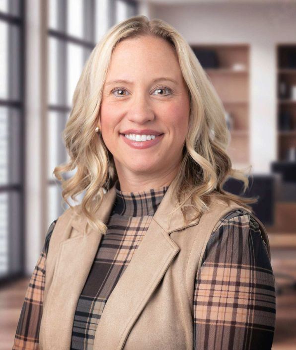 A professional headshot of a smiling SYM Financial Advisors team member in a plaid high-neck top and a tan vest, standing in an office setting; SYM Financial Advisors is an independent, commission-free fiduciary with 55+ years of serving clients.