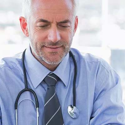 A middle-aged man with short gray hair and a beard is wearing a blue dress shirt and a striped tie. He has a stethoscope around his neck and is looking down with a slight smile, appearing focused. The background is blurred.