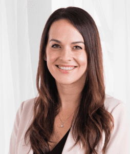 A woman with long, straight brown hair is smiling, standing in front of a light-colored background. She is wearing a light pink blazer with a black top underneath and a delicate necklace. The ambiance is bright and professional.