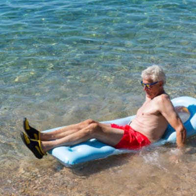 An elderly man with gray hair, wearing sunglasses, a red bathing suit, and black-and-yellow water shoes, relaxes on a light blue inflatable mattress at the edge of the clear, shallow sea water near a pebbled shore.
