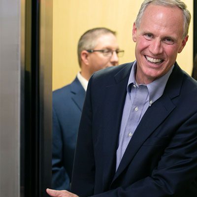 A man in a suit is smiling and holding an elevator door open. Another person in a suit is visible in the background, partially obscured. The background appears to be the interior of an elevator.