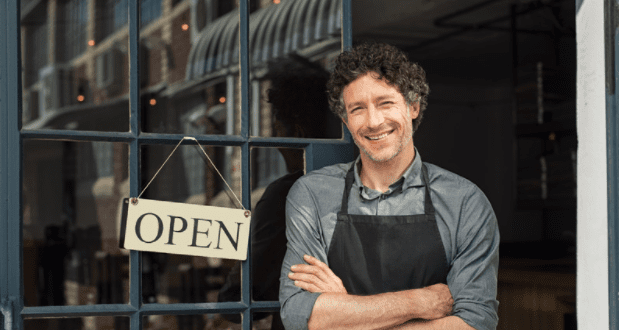 A smiling man with curly hair stands in front of a windowed door with an "OPEN" sign hanging on it. He is wearing a grey shirt and a black apron, arms folded, welcoming customers to his establishment. The interior and exterior of the shop are partially visible.