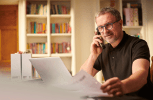 A man with glasses and a beard sits in front of a laptop in a home office with bookshelves in the background. He is smiling while talking on the phone and holding a sheet of paper. The ambiance suggests a relaxed yet productive work-from-home setting.