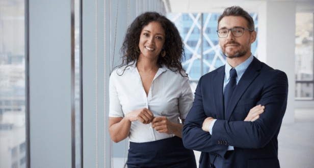 Two professionals stand in a modern office with large windows. The woman on the left has curly hair, wears a light-colored blouse and dark skirt, and has a relaxed smile. The man on the right has glasses, a beard, and wears a dark suit and tie with arms crossed.