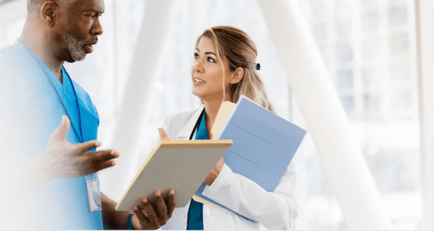 Two healthcare professionals, a man in blue scrubs and a woman in a white coat, are engaged in a discussion. The woman is holding a blue folder and a notepad, while the man is holding a yellow notepad. They appear to be in a modern, well-lit facility.