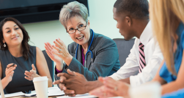 A diverse group of professionals are seated around a conference table engaged in a discussion. A woman with short gray hair and glasses is speaking and gestures with her hands. They have various documents and coffee cups in front of them.