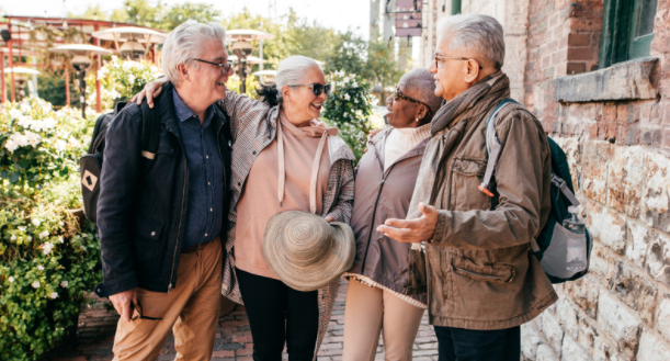 Four older adults, two men and two women, stand close together, smiling and chatting outdoors. They are dressed in casual, layered clothing and appear to be enjoying each other's company on a sunny day in a charming town setting with greenery and brick walls in the background.