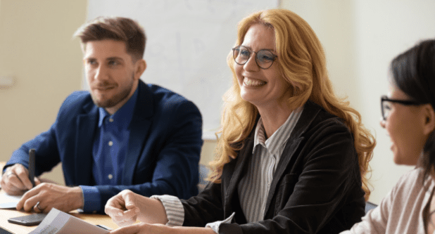 Three people are sitting at a conference table, engaged in discussion. The person in the middle, a woman with long blonde hair and glasses, is smiling. The man on the left is taking notes, while the woman on the right is looking at the blonde woman.