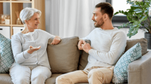 A senior woman and a younger man are sitting on a beige sofa in a cozy living room, likely discussing the joys and challenges of caring for aging parents. They are engaged in a friendly conversation, both smiling. The room features a white curtain, wooden shelves with various items, and a large green potted plant.